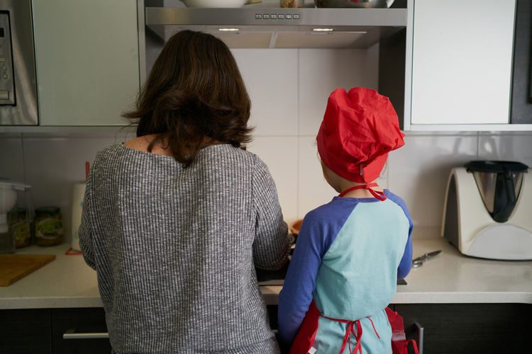 Backwards mother teaches daughter in red chef's hat in the kitchen