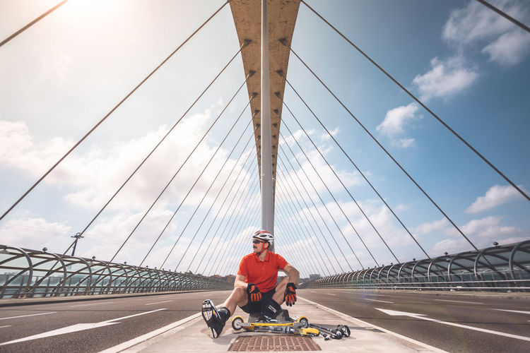 Man sitting with push scooter on bridge against cloudy sky