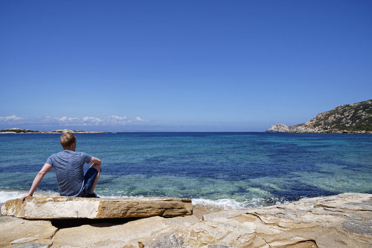 Rear view of woman sitting on rock by sea against clear blue sky