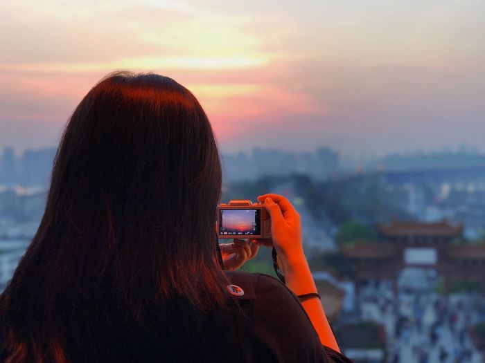 Rear view of woman photographing cityscape against sky during sunset