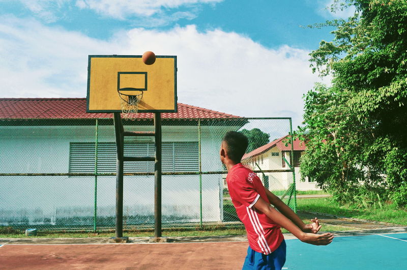 Teenage boy throwing ball in basketball hoop against trees