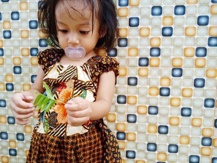 Cute girl sucking pacifier while holding plant against wall