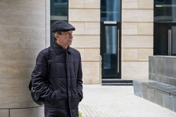 Adult caucasian male stands near modern building with columns. pensioner in seasonal clothes