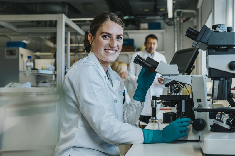 Smiling woman analyzing human brain slide under microscope while sitting with scientists in background at laboratory