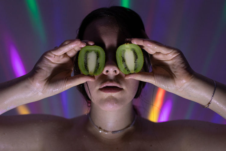 Close-up of woman covering eyes with kiwi slices against lights on wall