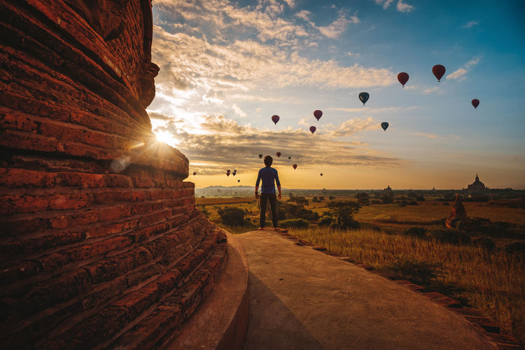 Rear view of man standing against hot air balloons flying in sky during sunset