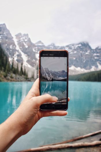 Hand holding mobile phone taking photo of lake and mountains