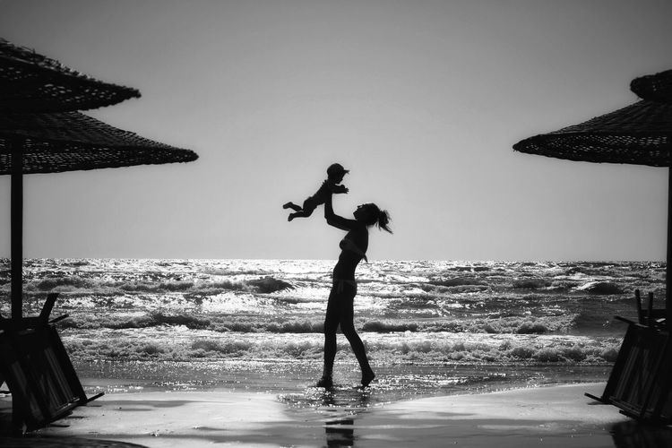 Mother standing on beach against clear sky over the sea playing with baby boy in her arms