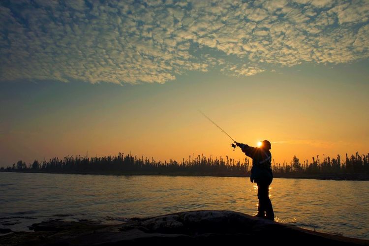 Silhouette man fishing in lake against orange sky