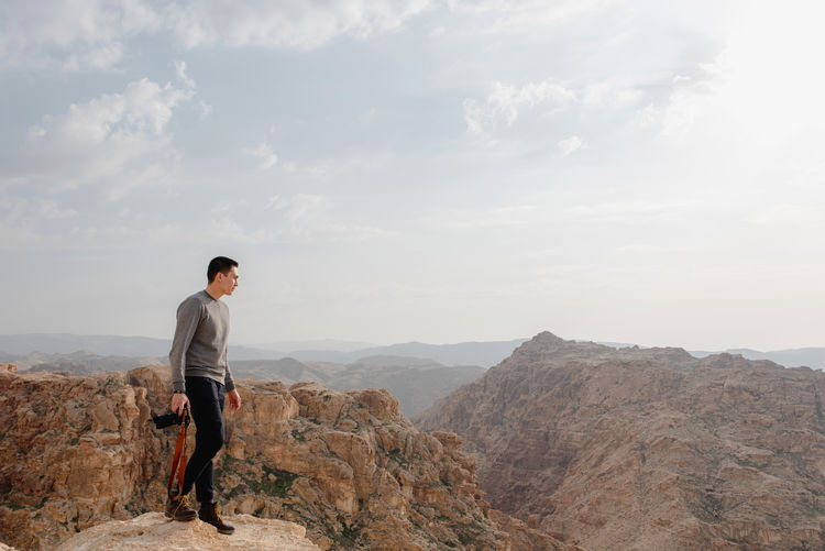 Side view of young stylish male photographer in trendy outfit admiring picturesque scenery of rocky mountains with camera in hand during trip in jordan