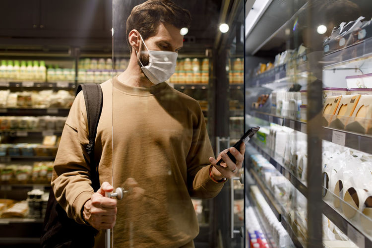 Male customer wearing protective face mask while using smart phone in supermarket during covid-19