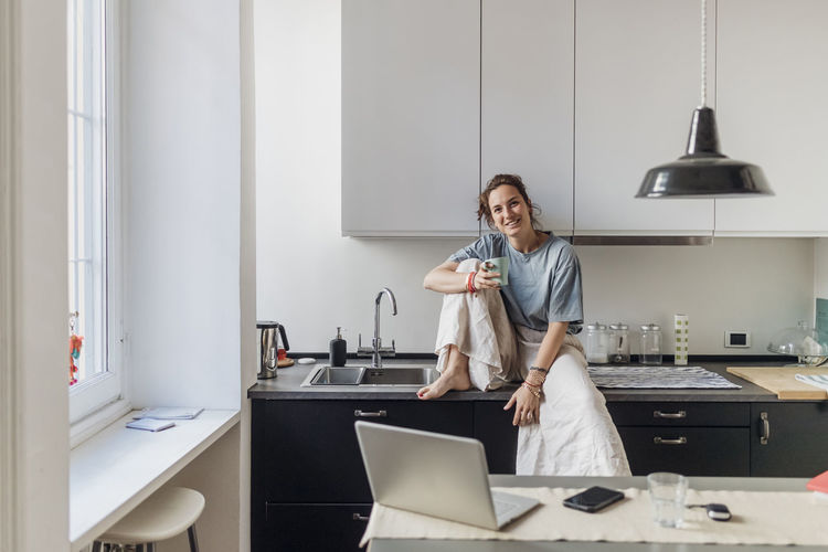 Smiling female freelancer holding coffee cup while sitting on kitchen counter