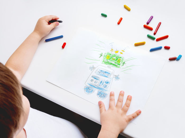 Left-handed toddler draws funny robot. kid uses wax crayons. top view on child's hands and pencils.