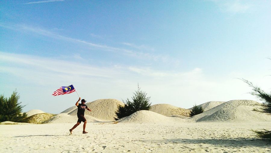 Mid adult man with malaysian flag running on sand against sky