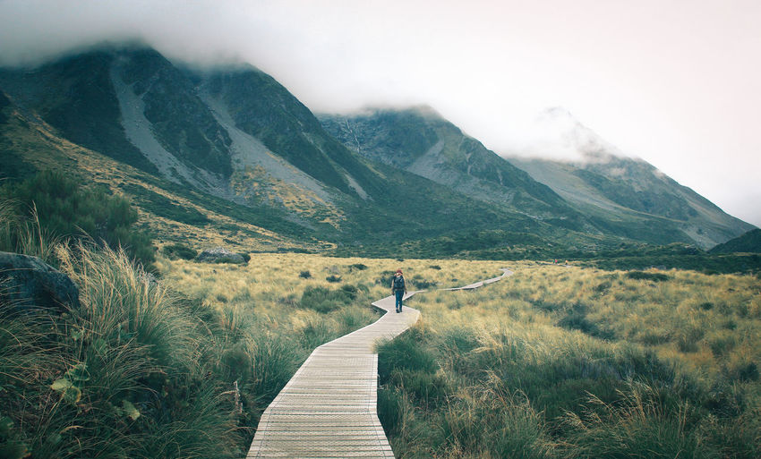 Woman walking on boardwalk leading towards mountains against sky