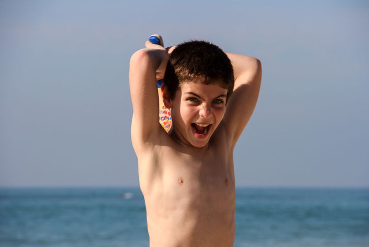 Shirtless boy holding baseball bat behind head at beach against sky