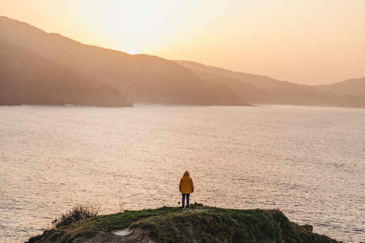 Back view of unrecognizable man in vibrant yellow jacket and denim standing on rocky hill and enjoying picturesque scenery of sea coast during sunset in spain