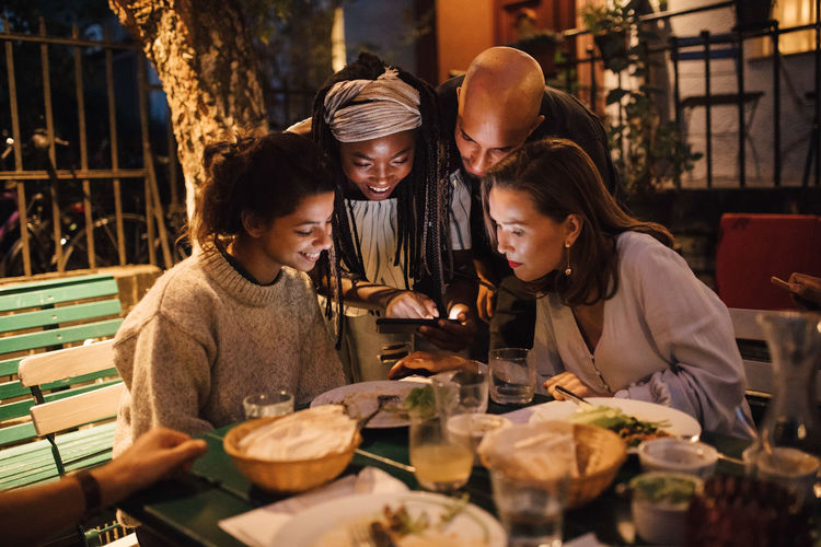Happy young woman showing smart phone to friends at table during dinner party