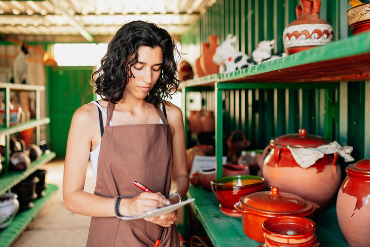 Young woman writing in note pad while standing by shelf in workshop