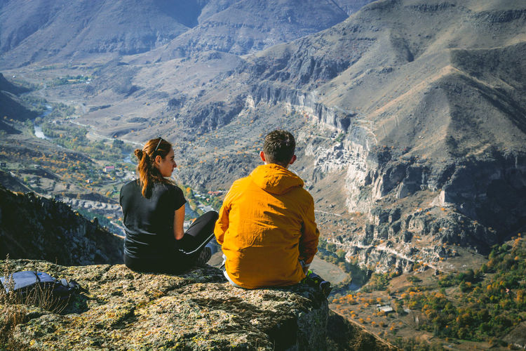 Rear view of couple sitting on cliff against mountains