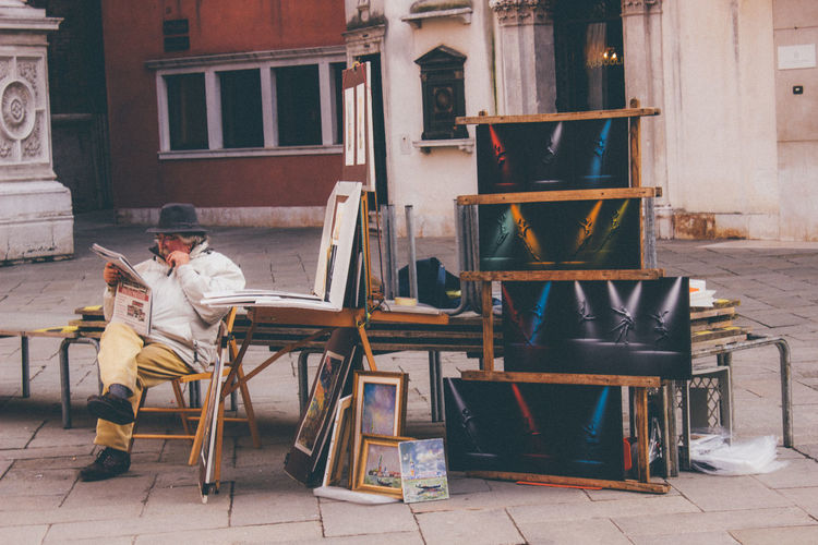 Man reading newspaper while selling paintings in city