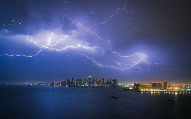 Lightning over illuminated city by sea at night