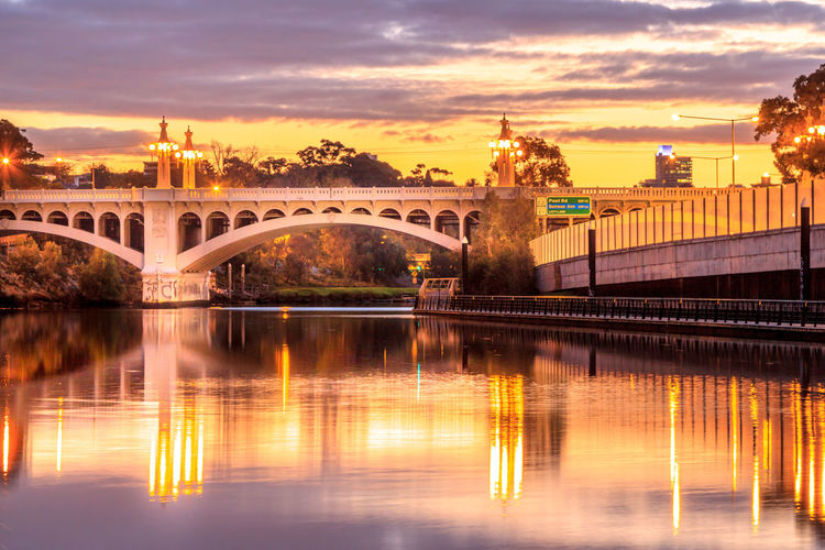 Illuminated arch bridge over river against cloudy sky during sunset