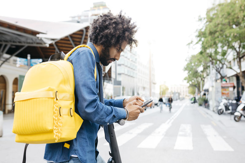 Man with backpack and e-scooter using navigation app on his mobile phone in the city, barcelona, spain
