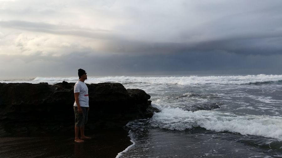 Side view of man standing at beach against cloudy sky
