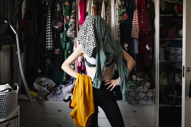 Dresses covering woman's face while standing with hands on hip against clothes rack in wardrobe