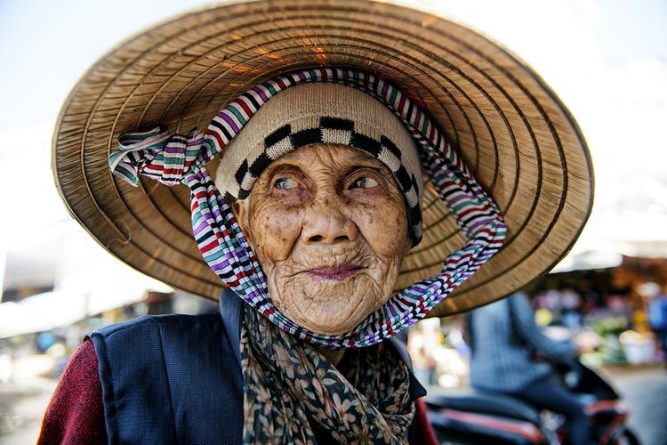 Wrinkled senior woman looking away while wearing asian style conical hat on street