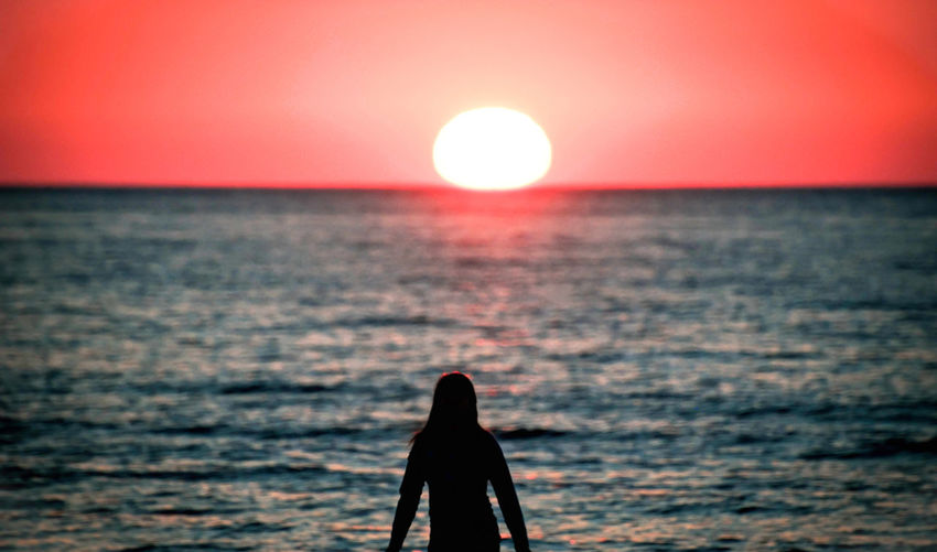 Silhouette of a young woman standing on the seashore and looking at a beautiful sunset.