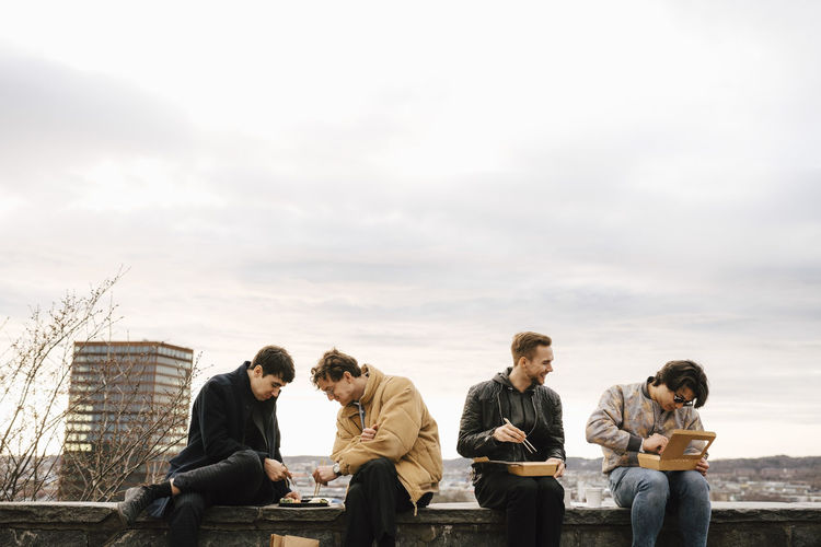 Multi-ethnic male friends eating package food while sitting on retaining wall against sky