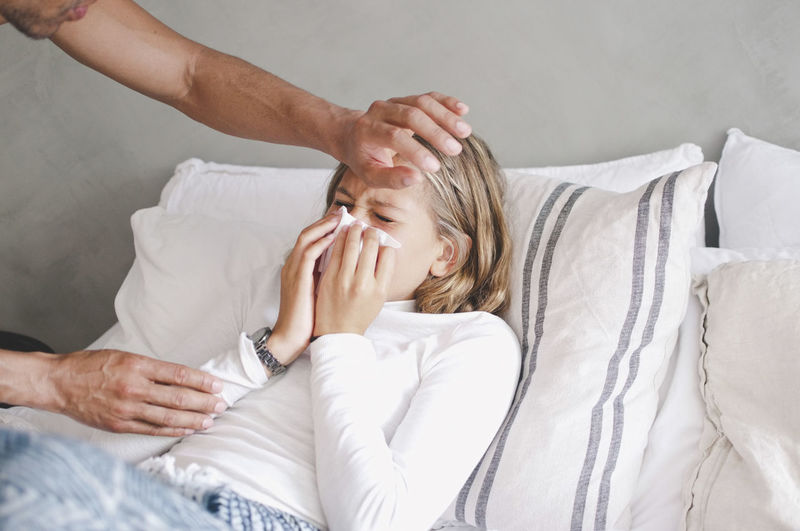 Father touching sick daughter blowing nose on bed at home