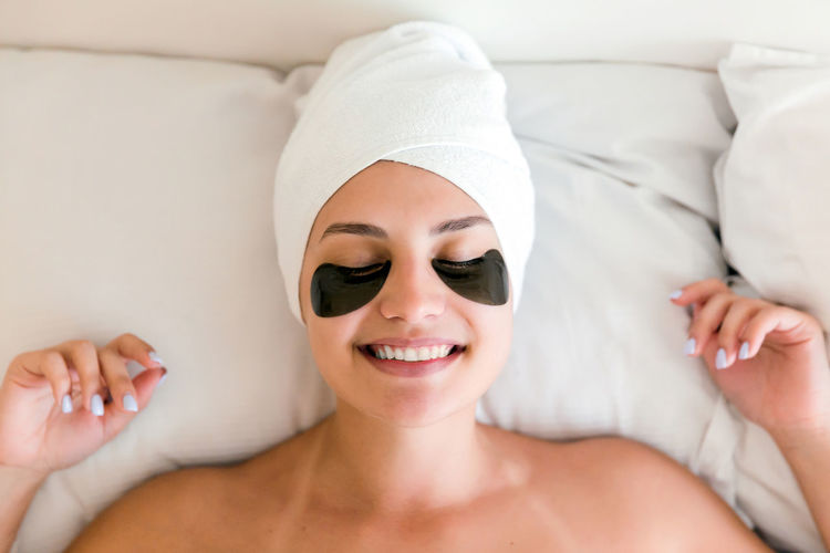 Smiling young woman with under eye patches lying on bed at spa