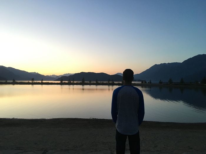 Rear view of man looking at lake against clear sky during sunset