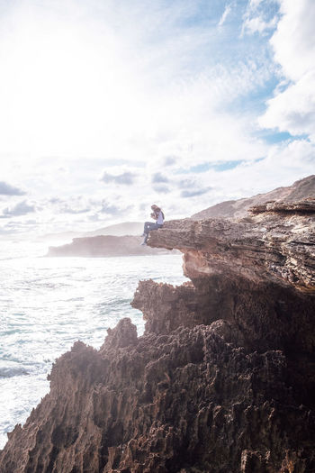 Woman sitting on cliff by sea against cloudy sky during sunny day