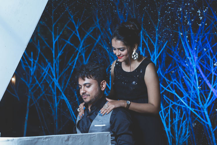 Young couple looking at camera at night against blue bushy background