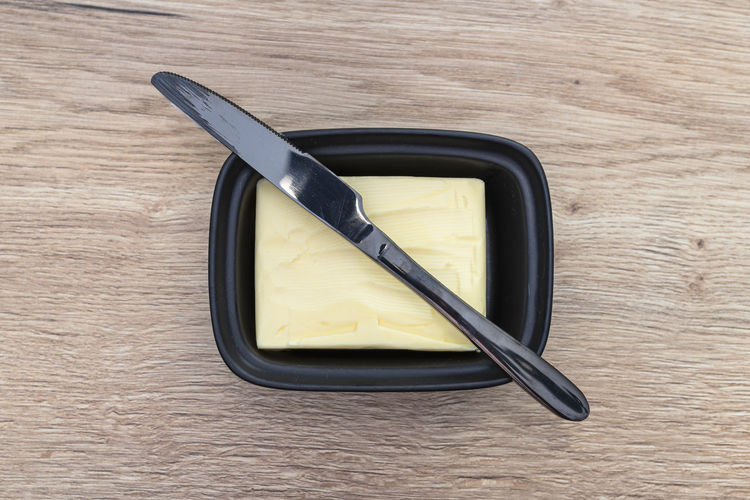 Black butter knife lying on a ceramic butter container, on a wooden table.