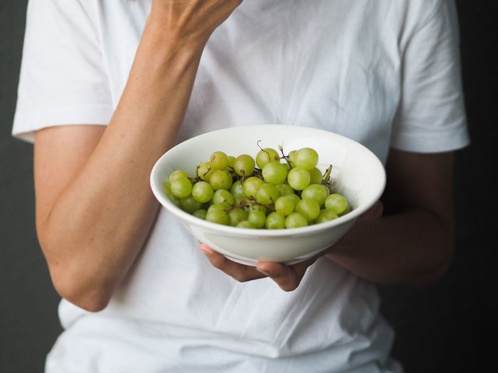 Unrecognisable woman wearing white t-shirt eating green grapes isolated on gray background. 