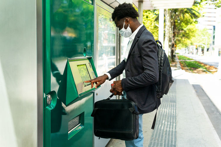 Side view of african american businessman in protective mask with suitcase using street atm terminal in barcelona city