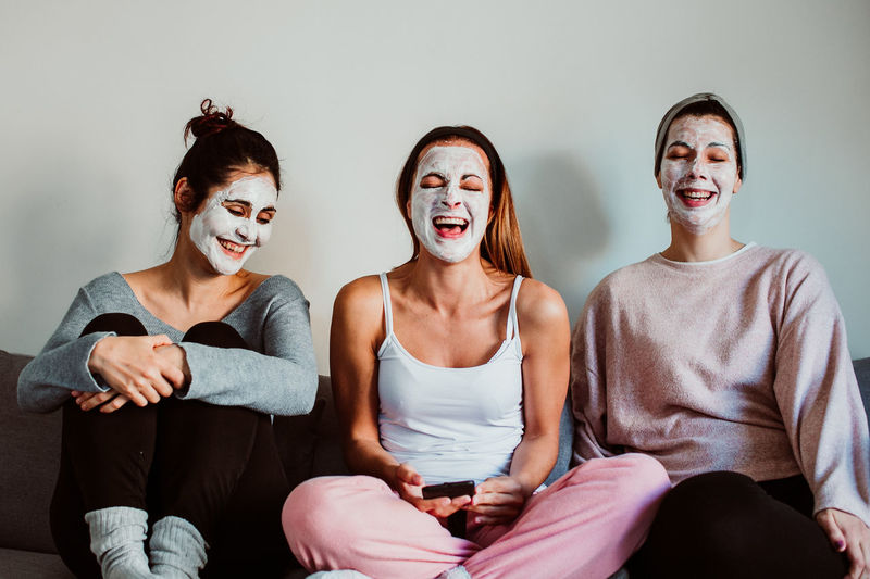 Cheerful female friends with facial masks sitting against wall at home