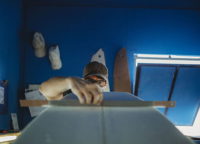 Concentrated qualified worker in protective mask aligning white surfboard in workshop with blue walls