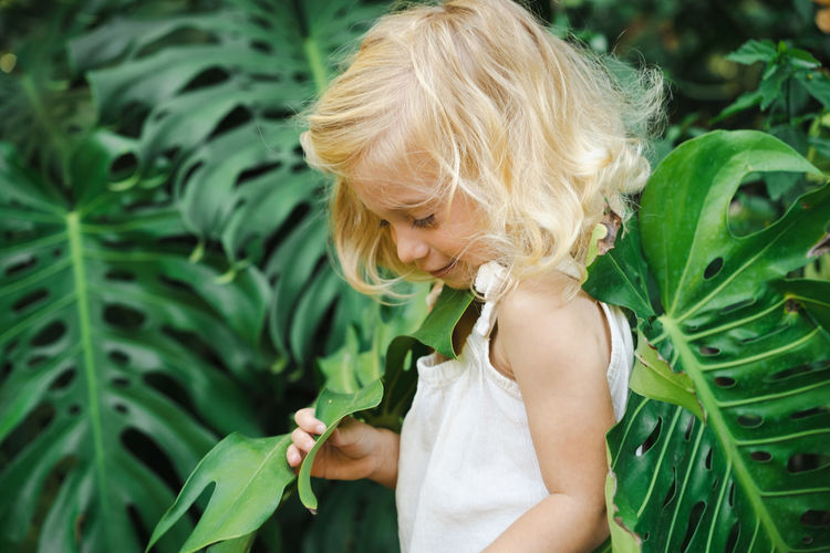 Cute beautiful blonde two year old girl, toddler, kid among big green exotic monstera leaves