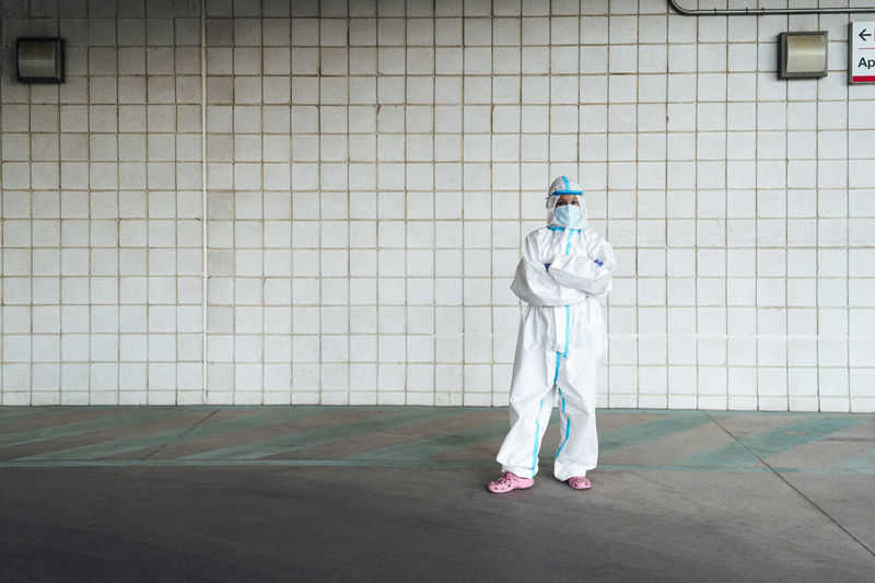 Female nurse wearing protective suit with arms crossed standing against tiled wall