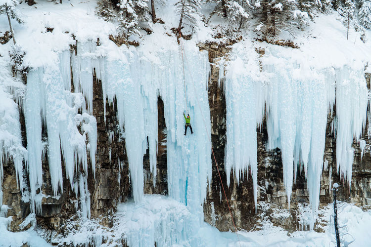 Panoramic view of snow-covered land and trees in forest with ice climber on frozen waterfall.