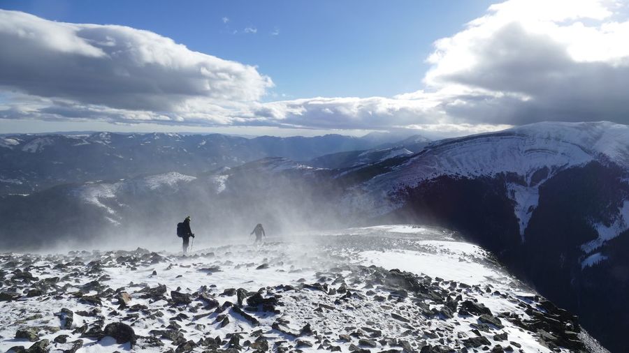 Hikers on a windy, snowy ridge with dramatic cloud.