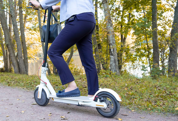 An adult elegant woman is about to ride an electric scooter in the park in early fall. 
