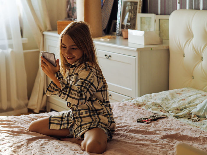 Cute girl using mobile phone sitting on bed at home