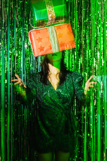 Focused female in green dress standing in studio with foil tinsel behind and tossing up christmas gift at party